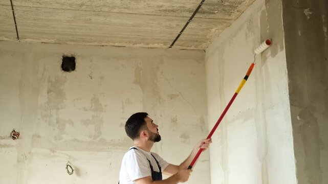 A bearded man in a work uniform is priming the walls after applying gypsum plaster. 