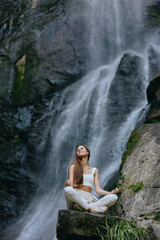 Obraz premium Woman meditating in lotus pose near a majestic waterfall, dressed in white fitness clothes, peaceful nature scene, calm and serene atmosphere, outdoor relaxation.