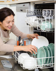 Smiling housewife putting dishes in dishwasher in kitchen at home