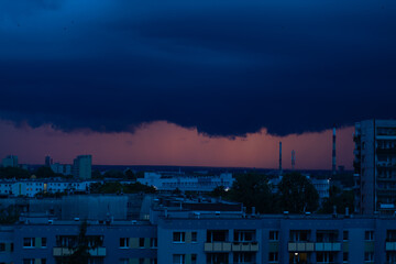 Dark storm clouds cover the city against the backdrop of a fiery sunset. Residential buildings and streets are plunged into shadow before the storm begins.