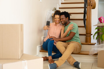 Happy young multiracial couple sitting on staircase while taking selfie at new home