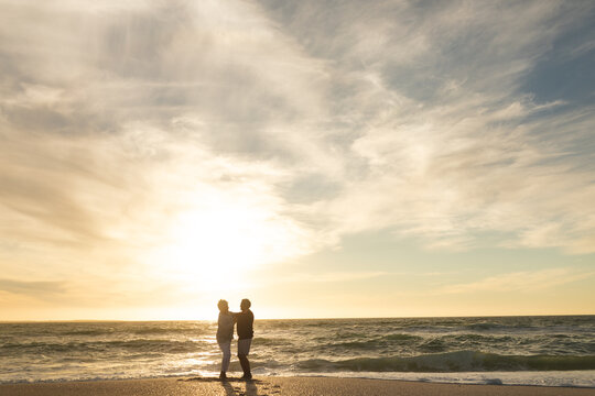 Full length of multiracial senior couple dancing on shore at beach against sky during sunset