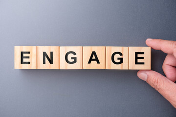 Wooden letter blocks spelling ENGAGE arranged by a human hand on a neutral background, representing engagement, interaction, and connection concept for business, marketing, and communication