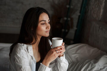A woman with black hair wakes up in the morning in a white blanket and catches the rays of the sun. The portrait is creative, with brown eyes. High quality photo
