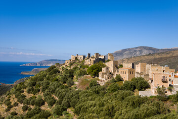 Fototapeta premium Traditional stone houses and towers sit atop a green hillside with panoramic views of the Mediterranean Sea near Vathia, Greece. The scene features rugged textures, sunlit architecture, and vibrant