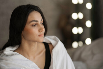 A woman with black hair wakes up in the morning in a white blanket and catches the rays of the sun. The portrait is creative, with brown eyes. High quality photo