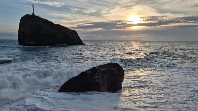 Sea sunset rocks, waves crash on large rock formations with a distant island and cross, a peaceful ocean view