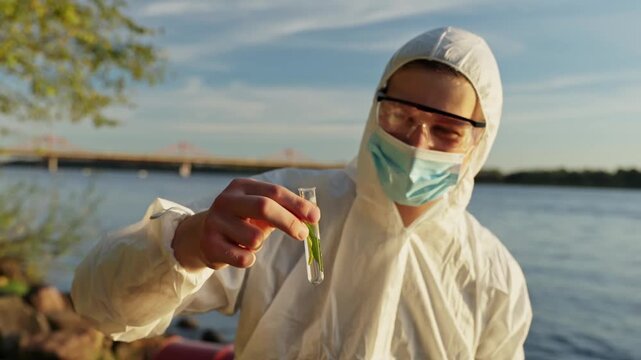 Masked scientist in white hazmat suit holds a vial with green water sample by a river at sunset, checking pollution levels and documenting results near a bridge.