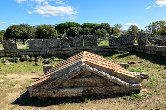 A weathered stone tomb with a triangular roof sits among ancient masonry blocks at the archaeological site of Paestum, Italy. The scene is set under crisp sunlight with lush green trees and a vivid