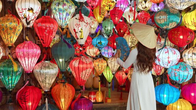 4K slow-motion video shows an Asian woman wearing a white Vietnamese traditional dress and conical hat, holding a traditional fan, amidst colorful lanterns in Hoi An, Da Nang, Vietnam.