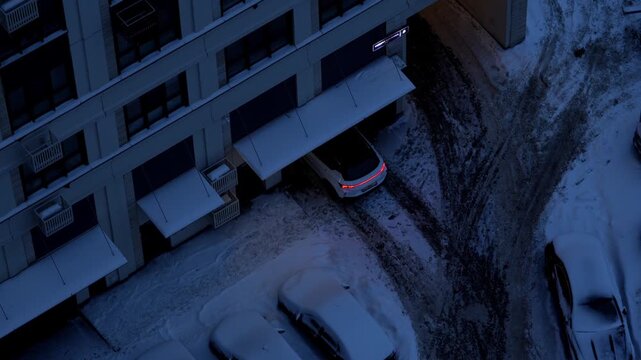 Car entering parking of a residential building during harsh winter weather with snow, cold temperatures, and quiet evening atmosphere, representing difficult seasonal conditions and everyday urban rea