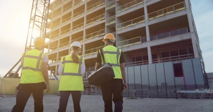 Three Construction Professionals review Digital Plans on a Tablet While Walking Across Site, Engineering Tasks for Commercial and Residential High‑rise Development with Under Construction Facade.