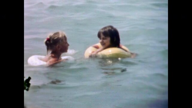 Girls friends swim in inflatable rings in sea on sunny summer holiday. Children chat, laugh together. Happy childhood friendship in seaside leisure. Old film. Archival retro 1970s. Vintage archive