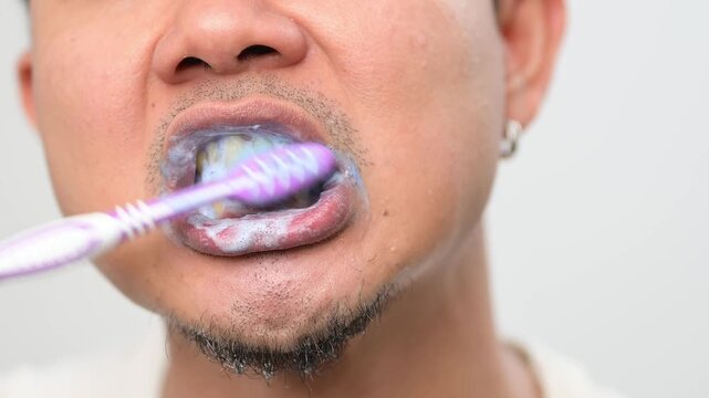 Close up video of an Asian adult man brushing his teeth with purple toothpaste foam. Oral hygiene routine concept for brighter looking teeth, dental care and personal healthcare.