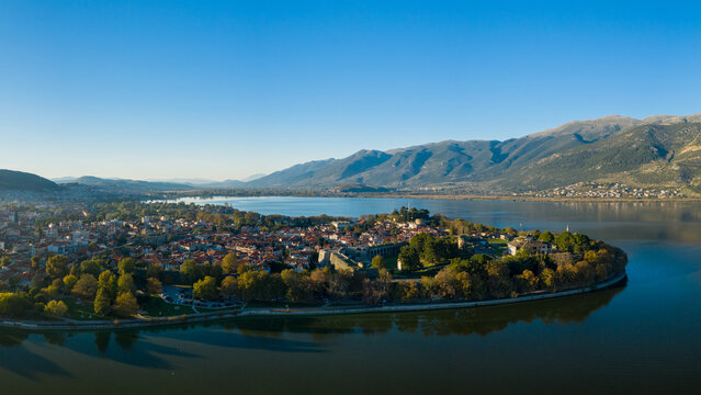 Soft sunlight illuminates the historic peninsula of Ioannina, surrounded by the calm waters of Lake Ioannina and backed by the rugged slopes of the Pindus mountains. The image features red-tiled