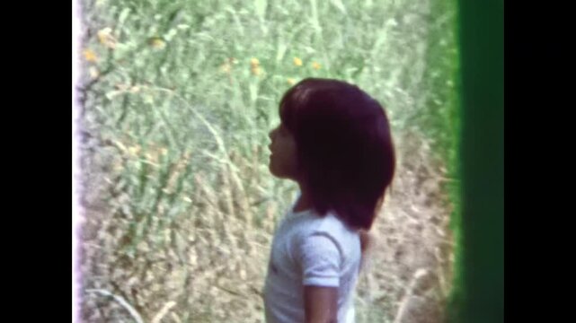 Girl child smelling wildflowers in sunny summer meadow. Child enjoying fresh, green nature outdoors. Peaceful childhood activity in countryside field. Old film. Archival retro 1970s. Vintage archive