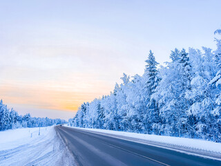 Winter road through a snow-covered forest at sunset, scenic asphalt highway in a cold frosty morning with trees in white hoarfrost