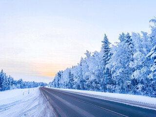 Winter road through a snow-covered forest at sunset, scenic asphalt highway in a cold frosty morning with trees in white hoarfrost