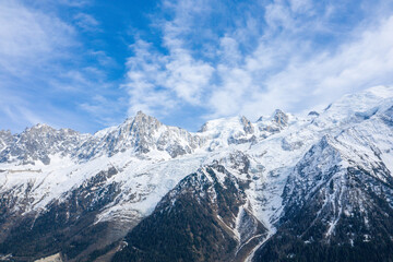 Fototapeta premium Sharp, rugged summits of the Mont Blanc massif rise above a glacier and dark forested slopes, with textured clouds drifting across a bright blue sky.