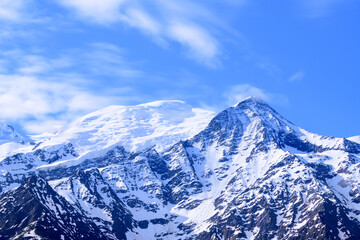 Sunlit snowy slopes of Mont Blanc and Aiguille du Gouter stand beneath a vibrant blue sky, with soft clouds drifting above the rugged alpine peaks. © Florent