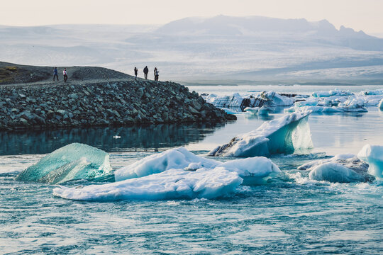 View of serene icebergs drift on a calm glacial lagoon, reflecting the soft light under a hazy sky, with distant figures on a rocky shore, Hofn, Vatnajokull, Iceland.
