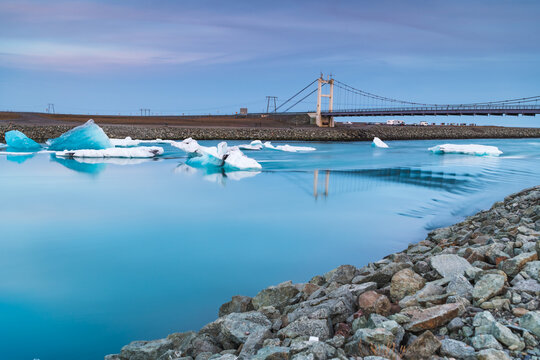 View of luminous blue icebergs drift serenely in the glacial lagoon beneath a modern bridge against the backdrop of a tranquil twilight sky, Hofn, Vatnajokull, Iceland.