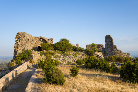 Weathered stone walls of Chateau de Opoul Perillos stand atop a rugged hill, surrounded by wild grasses and shrubs in warm afternoon light under a clear blue sky.