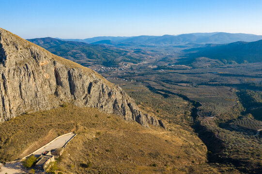 Aerial view of dramatic rocky cliffs on Acrocorinth rising above sunlit plains and patchwork farmland, with distant mountains under a clear blue sky in Greece.