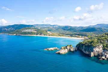 Obraz premium Expansive view of Karavostasi beach in Epirus, Greece, with turquoise sea, sandy shoreline, and rugged rocky outcrops surrounded by lush green hills. Bright daylight and scattered clouds enhance the