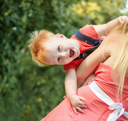 Closeup portrait of laughing baby boy. Mom and son are playing together in the park in the summer. Mother is holding kid in her arms.