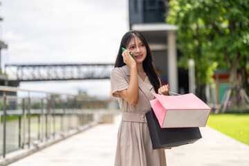 Fototapeta premium Happy asian woman holding shopping bags talking on phone about credit card discount, Girl shopper enjoying end of season sale promotion at department store, Mobile app delivery lifestyle concept