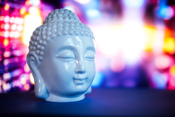 White ceramic Buddha head sculpture with serene expression, placed on a dark surface, surrounded by colorful bokeh lights, creating a tranquil and meditative atmosphere