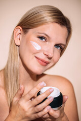 Blonde woman applying skincare cream to her face while holding a jar of moisturizer, showcasing beauty routine and self-care practices in a serene environment
