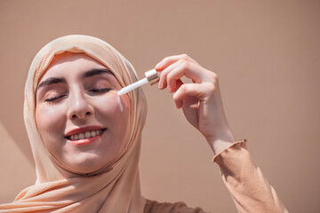 Young woman with light skin and hijab applying serum to her face, showcasing skincare routine, with soft lighting and neutral background emphasizing beauty and self-care