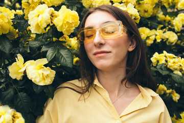Young woman with long brown hair, wearing yellow sunglasses, poses among vibrant yellow flowers, creating a cheerful and lively atmosphere in a natural setting