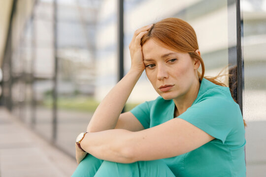 Healthcare worker sitting outside a hospital, feeling tired and stressed, leaning on a glass wall.