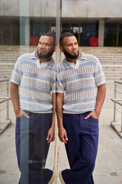 Young man standing outdoors reflecting against glass in urban setting