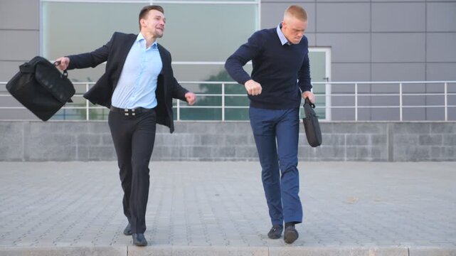 Two happy young businessmen with briefcases funny dancing while walking after successful business deal outdoor. Cheerful handsome colleagues celebrating their achievement near office center. Close up