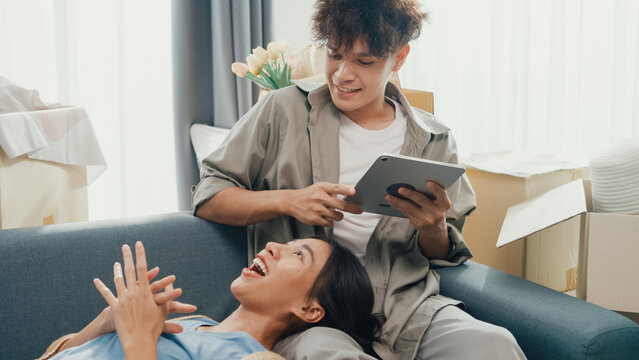 Young happy Asia couple sit on sofa couch use digital tablet online shopping furniture from decoration store in living room at home. Husband wife move new house, Apartment Rent And Ownership Concept. - Powered by Adobe