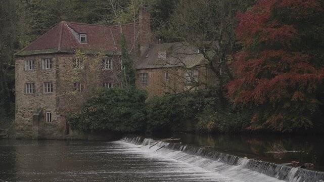A static medium shot focusing on the historic stone Fulling Mill on the banks of the River Wear, with water cascading over the famous weir in the foreground on a moody autumn morning in Durham.