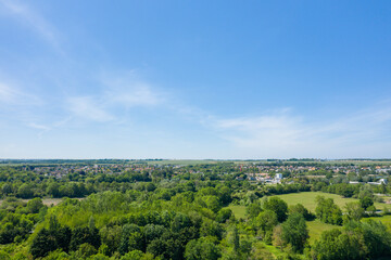Obraz premium Wide aerial view of lush green fields and dense forest leading to a distant village under a clear blue sky in Normandy.