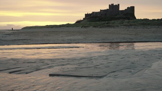 A low-angle static wide shot of the majestic Bamburgh Castle silhouetted against a golden sunrise sky, viewed from the sandy beach with a shallow stream reflecting the morning light in the foreground.
