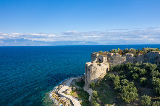 A weathered stone bastion stands above the vivid blue Mediterranean, surrounded by olive trees and set against a backdrop of distant mountains and dramatic sky near Koroni, Greece.