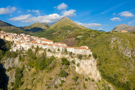Picturesque view of Tortora, Italy, with white stone houses and terracotta roofs perched atop a steep cliff, surrounded by lush green hills and rugged mountains under a bright blue sky.