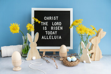 Phrase CHRIST THE LORD IS RISEN TODAY ALLELUIA traditional greeting to christian holiday. Festive mood composition with bunnies and yellow flowers on pastel blue background. Sustainable natural