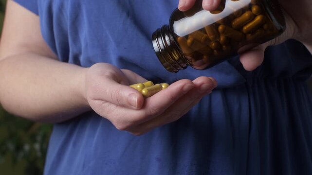 Closeup woman pours supplement capsules from amber bottle into palm indoors, measuring daily dose for selfcare routine today.