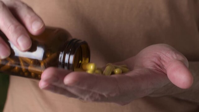 Adult man pours dietary supplement capsules into palm from amber bottle, close up at home to measure daily dose for wellness.