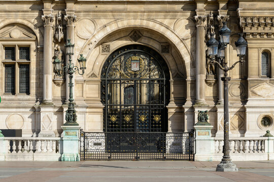 Elegant arched iron gate framed by classical stone columns and decorative lanterns on the facade of Hotel de Ville in Paris. Sunlight reveals intricate metalwork, sculpted details, and historic