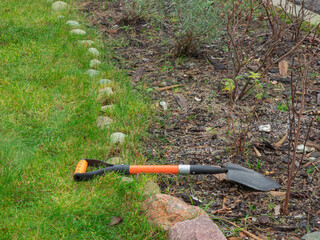 A small orange and black spade is on a green lawn and a mulched garden bed, symbolizing gardening work, home improvement, and preparing for the planting season