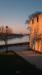 Riverside promenade with historic stone castle wall bathed in golden hour light, an old lamppost glowing by the calm river and distant city skyline, serene and nostalgic